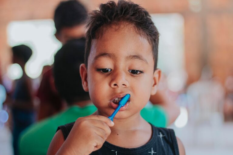 A young child brushes their teeth at a bathroom sink while an adult supervises with a smile.